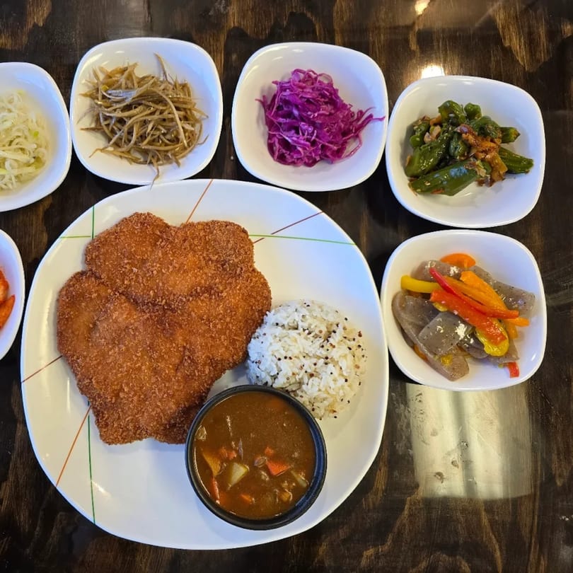 Korean tonkatsu with curry, rice, and various banchan side dishes on a wooden table.