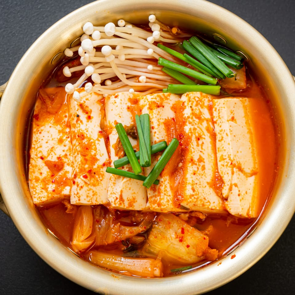 Overhead view of a golden pot of Korean kimchi stew with tofu, mushrooms, and green onions.