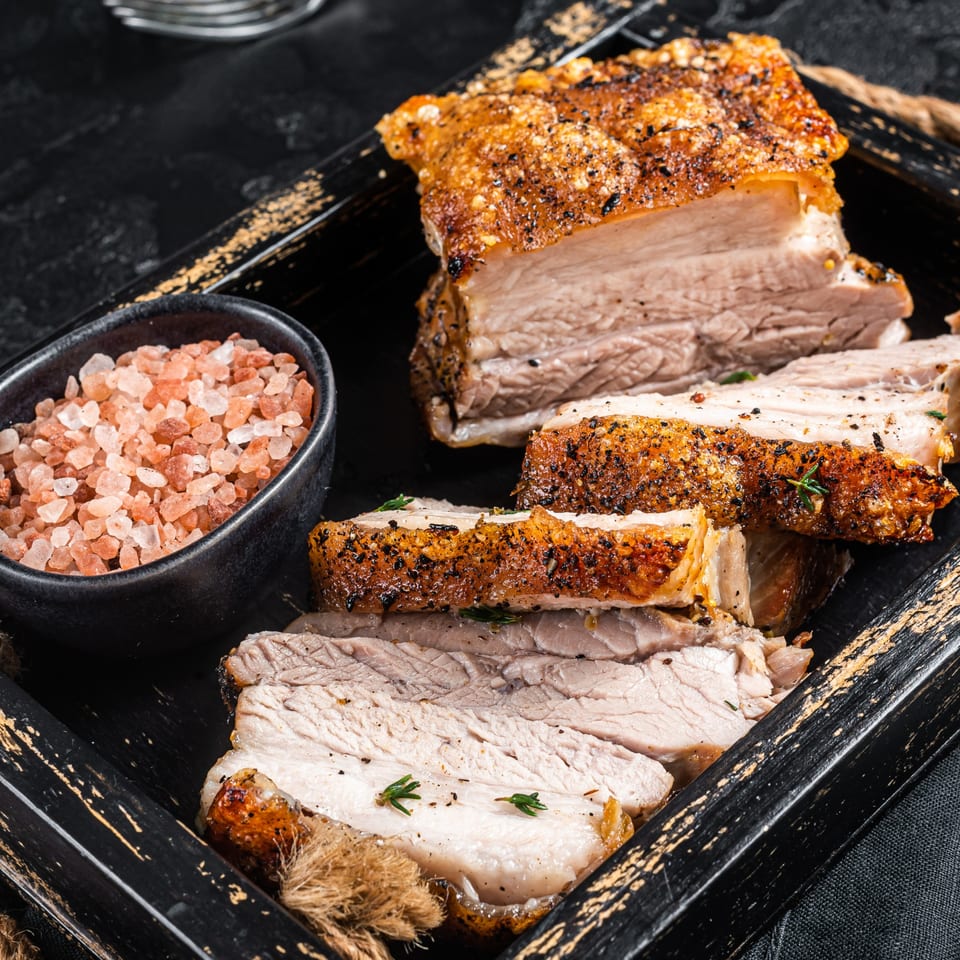 Crispy roasted pork belly slices with pink Himalayan salt in a black wooden tray.