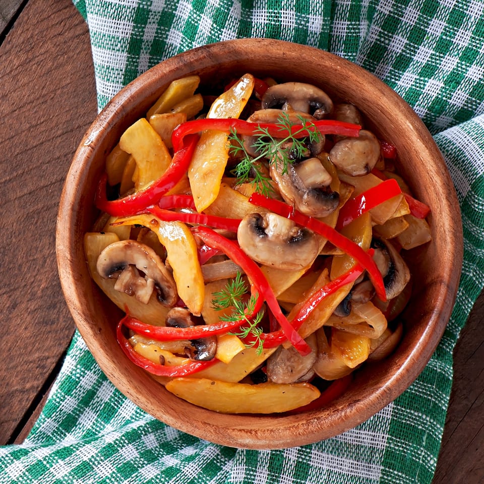 A wooden bowl filled with Korean-style fried potatoes, red peppers, mushrooms, and dill on a green checkered cloth.