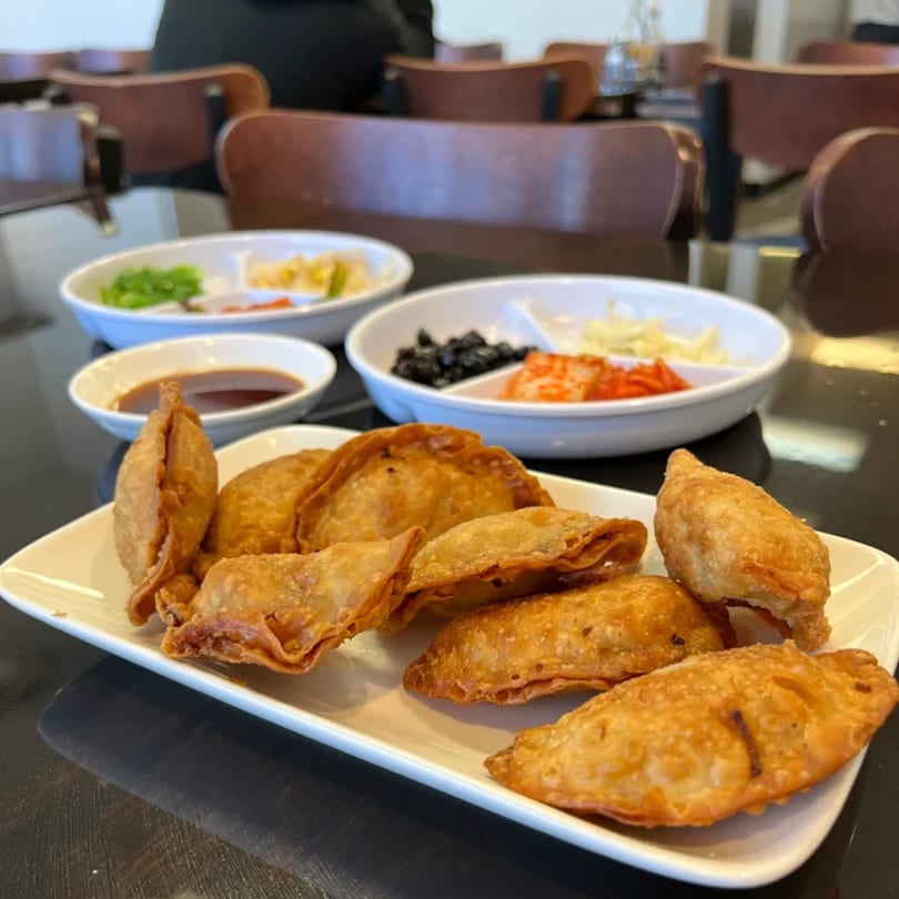 A plate of golden-brown Korean fried dumplings with dipping sauce and banchan in the background.