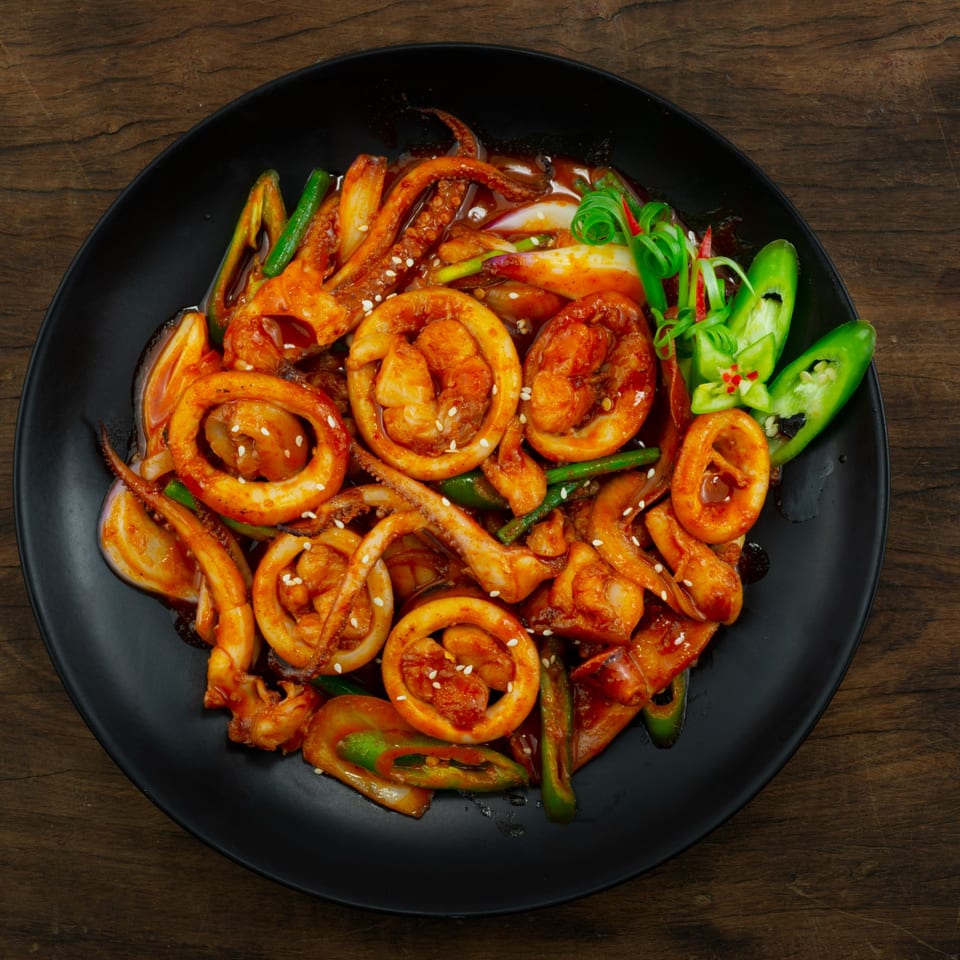 Spicy Korean stir-fried squid and shrimp with green onions and peppers on a black plate.