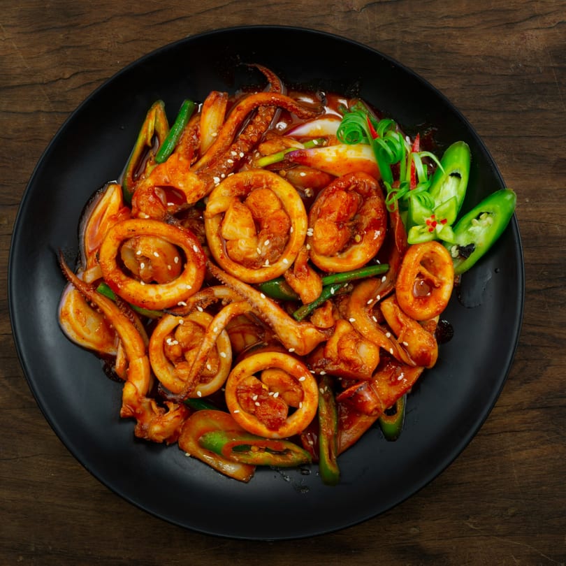 Spicy Korean stir-fried squid and shrimp with green onions and peppers on a black plate.